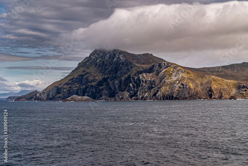 Cape Horn, Wollaston Islands, Chile - December 14, 2008: Dark rock with white and yellow patches mountain rises sharply out of  dark gray ocean and put its top in cloudscape with light blue patches. photo