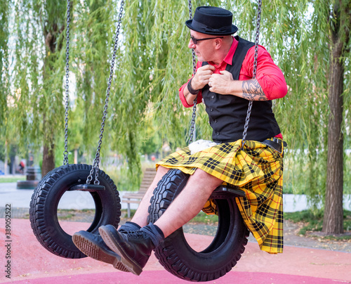 portrait of an eccentric man dressed in a kilt playing on swings made of tires, adult in child's attitude in a playground, immaturity concept