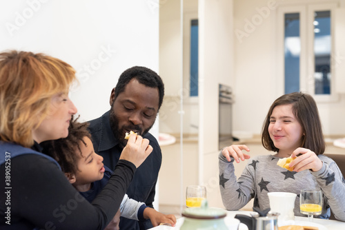 Happy interracial family with young children having breakfast in the kitchen, mixed-race family having fun at the table, moments of tenderness between the parents while the children eat happily