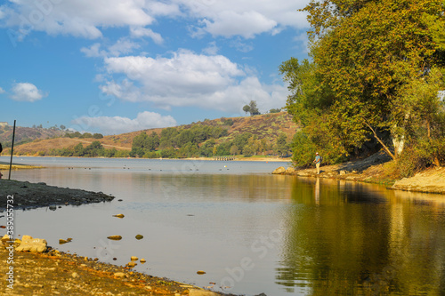 a gorgeous shot of the vast still lake water with lush green and autumn colored trees reflecting off the lake with people fishing on the banks of the lake at Puddingstone Lake in San Dimas California photo