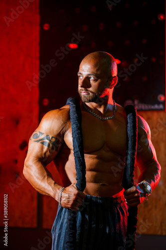 Sporty handsome strong man posing with sports rope on the background of the gym. A strong bodybuilder with perfect abs, shoulders, biceps, triceps and chest.