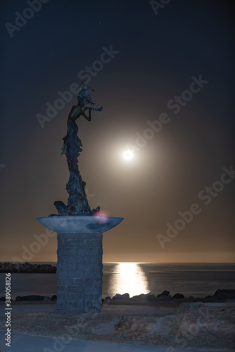 Mermaid statue stands lit against the full moon as it illuminates the ocean as it drops toward the horizon. photo