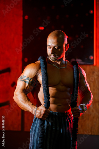 Sporty handsome strong man posing with sports rope on the background of the gym. A strong bodybuilder with perfect abs, shoulders, biceps, triceps and chest.