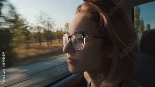 A girl with glasses rides in the back seat and looks out the open window.