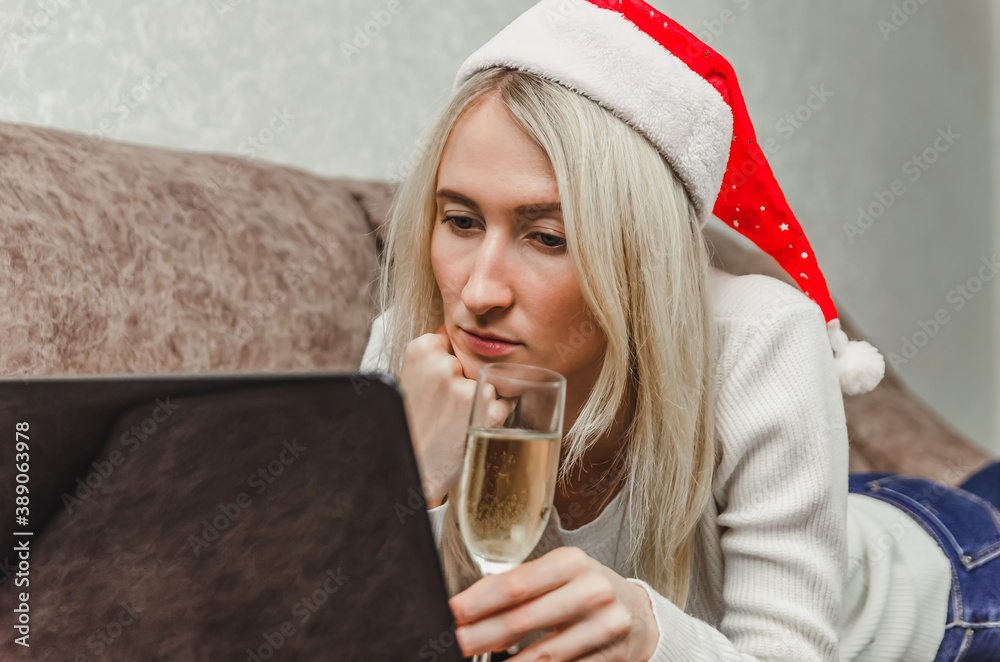 Young sad blonde woman in a Santa hat with a glass of champagne is lying on the sofa and communicating via a laptop video link. Concept of Christmas, New year, online communication.