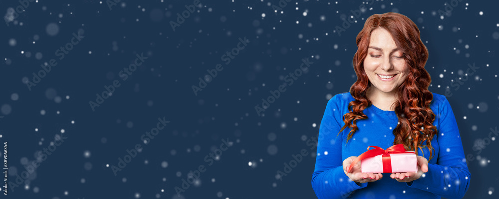 Cheerful woman celebrates christmas and new year. Young girl with curly hair holding a gift box with a red ribbon on a blue background. Snowflakes decor Cheerful woman celebrates christmas and new year. Young girl with curly hair holding a gift box with a red ribbon on a blue background. Snowflakes decor