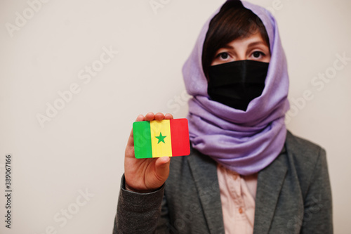 Portrait of young muslim woman wearing formal wear, protect face mask and hijab head scarf, hold Senegal flag card against isolated background. Coronavirus country concept. photo