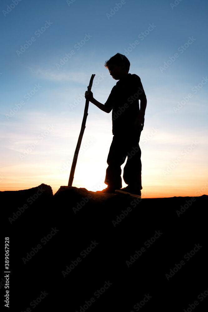 A young boy with a hiking stick silouetted over a sunset sky.
