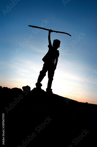 A young boy with a hiking stick silouetted over a sunset sky.