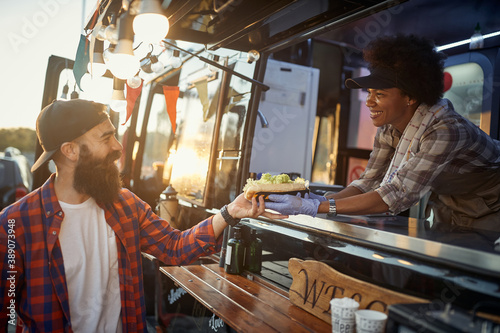 afro-american female employee gives with care sandwich to young hipster male customer photo