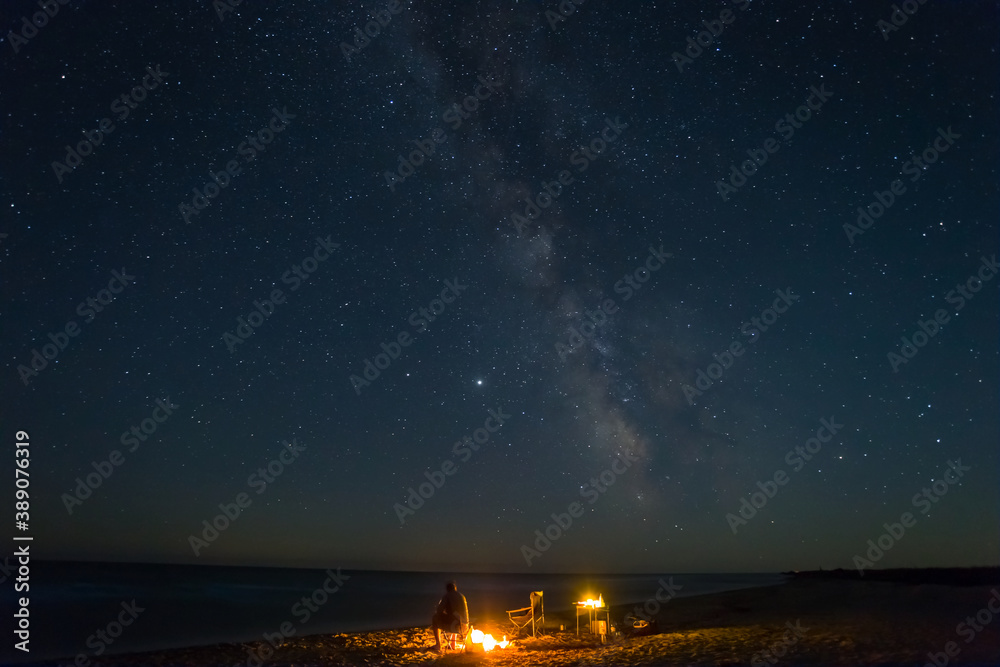 man sit on a chair near a fire on a sea coast under starry sky with milky way, night travel camping scene