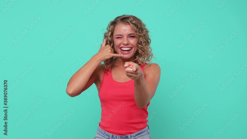 Cheerful young woman 20s years old in pink tank top posing isolated on blue turquoise background studio. People lifestyle concept. Doing phone gesture says call me back blinking point finger on camera Cheerful young woman 20s years old in pink tank top posing isolated on blue turquoise background studio. People lifestyle concept. Doing phone gesture says call me back blinking point finger on camera