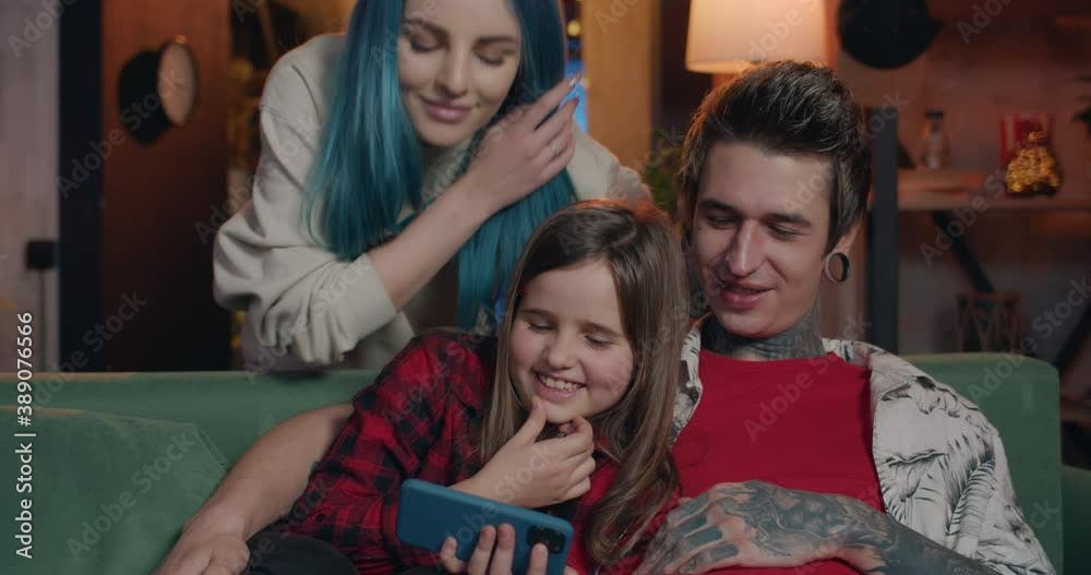 Millennial father with tattoos and piercing and his teen daughter looking on phone screen and laughing while coming mother. Portrait of modern family spending time together. Millennial father with tattoos and piercing and his teen daughter looking on phone screen and laughing while coming mother. Portrait of modern family spending time together.