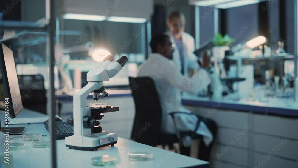 Shifting Focus from a Digital Microscope to Female and Male Medical Research Scientists Having Conversation While Conducting Experiments, Writing Results on a Tablet in Modern Science Laboratory. Shifting Focus from a Digital Microscope to Female and Male Medical Research Scientists Having Conversation While Conducting Experiments, Writing Results on a Tablet in Modern Science Laboratory.