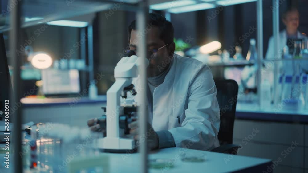Medical Research Scientist Conducts DNA Experiments Under a Digital Microscope in a Biological Applied Science Laboratory. Multiethnic Lab Engineer in Glasses in White Coat Working on Vaccine. Medical Research Scientist Conducts DNA Experiments Under a Digital Microscope in a Biological Applied Science Laboratory. Multiethnic Lab Engineer in Glasses in White Coat Working on Vaccine.