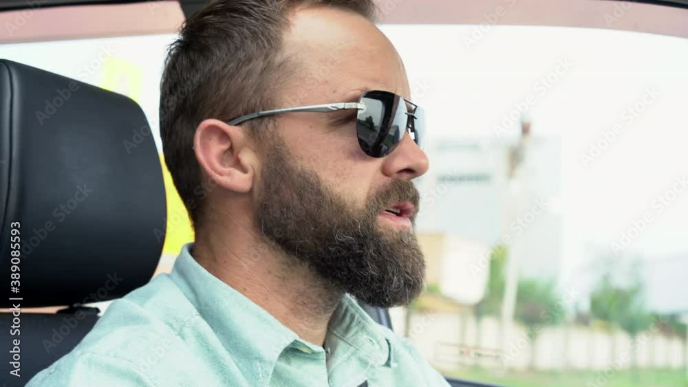 Adult bearded man in sunglasses driving the car. Young hipster drives a car and talks to a fellow traveler. Business man drives a vehicle. 
