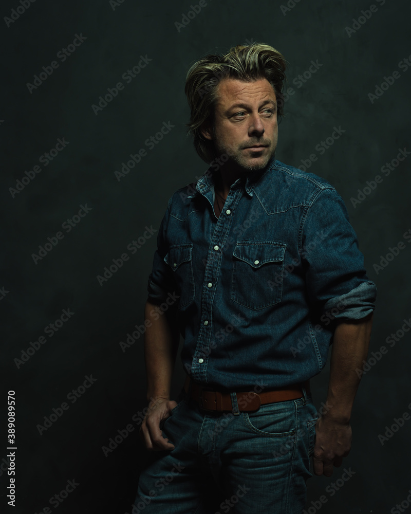 Atmospheric dark studio portrait of a tough middle-aged man with blonde hair in a jeans shirt. Atmospheric dark studio portrait of a tough middle-aged man with blonde hair in a jeans shirt.