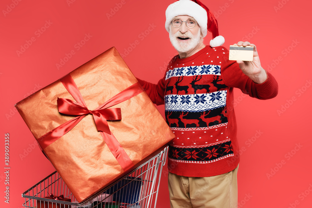 Smiling elderly man in santa hat holding credit card near gifts in shopping cart on red background