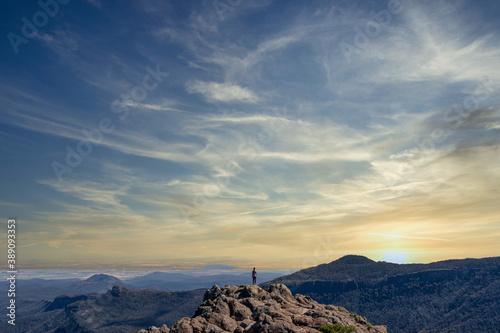 A young girl stands on top of a rock hill and looks at the sunset in a mountainous landscape.