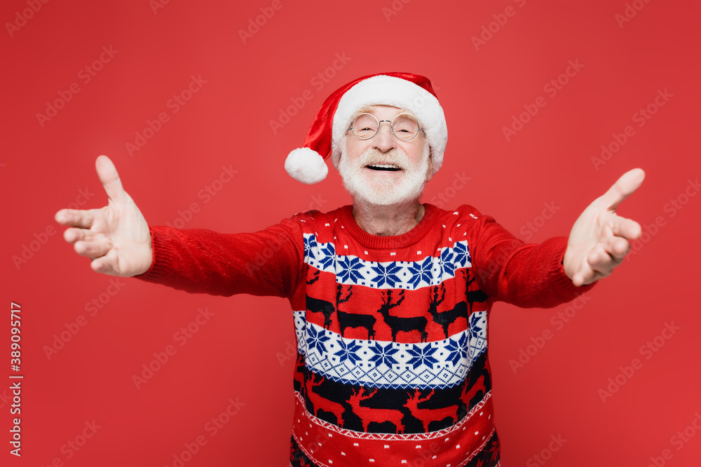 Smiling senior man in santa hat pulling hands at camera on blurred foreground isolated on red