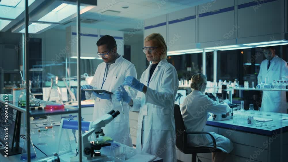 Female and Male Medical Research Scientists Have a Conversation While Conducting Experiments in Sample Test Tubes, Writing Analysis Results on a Tablet Computer. Modern Biological Science Laboratory. Female and Male Medical Research Scientists Have a Conversation While Conducting Experiments in Sample Test Tubes, Writing Analysis Results on a Tablet Computer. Modern Biological Science Laboratory.