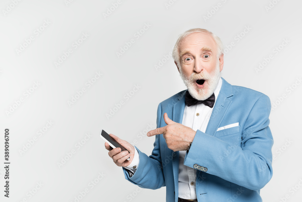 Excited senior man in bow tie and jacket pointing with finger at smartphone isolated on grey Excited senior man in bow tie and jacket pointing with finger at smartphone isolated on grey