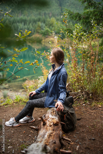 Close-up side view of beautiful tourist woman with backpack siting near azure lake on wooded mountain background. Amazing natural landscape. Concept of traveler.