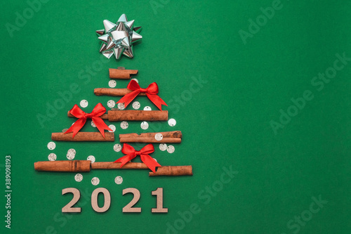 New year card 2021. Christmas tree made of cinnamon sticks with silver gift bow and small red bows on green background with silver confetti, copy space. Flat lay. template for design.