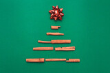 Christmas tree made of cinnamon sticks with a red gift bow on a green background, copy space. Flat lay. New year card and template for design.