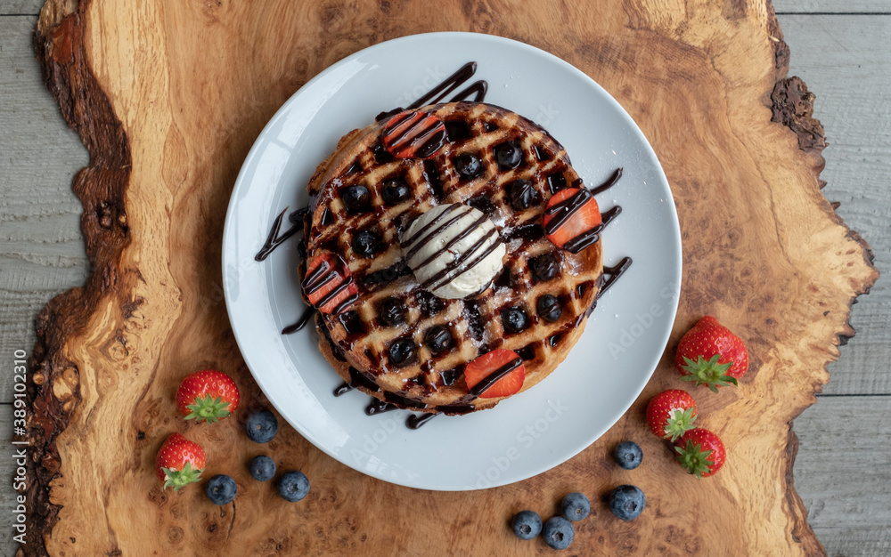 Fototapeta premium Vegan waffle with vanilla icecream, chocolate sauce, and decorated with blueberries and strawberries. Photographed from above on an olive wood platter.