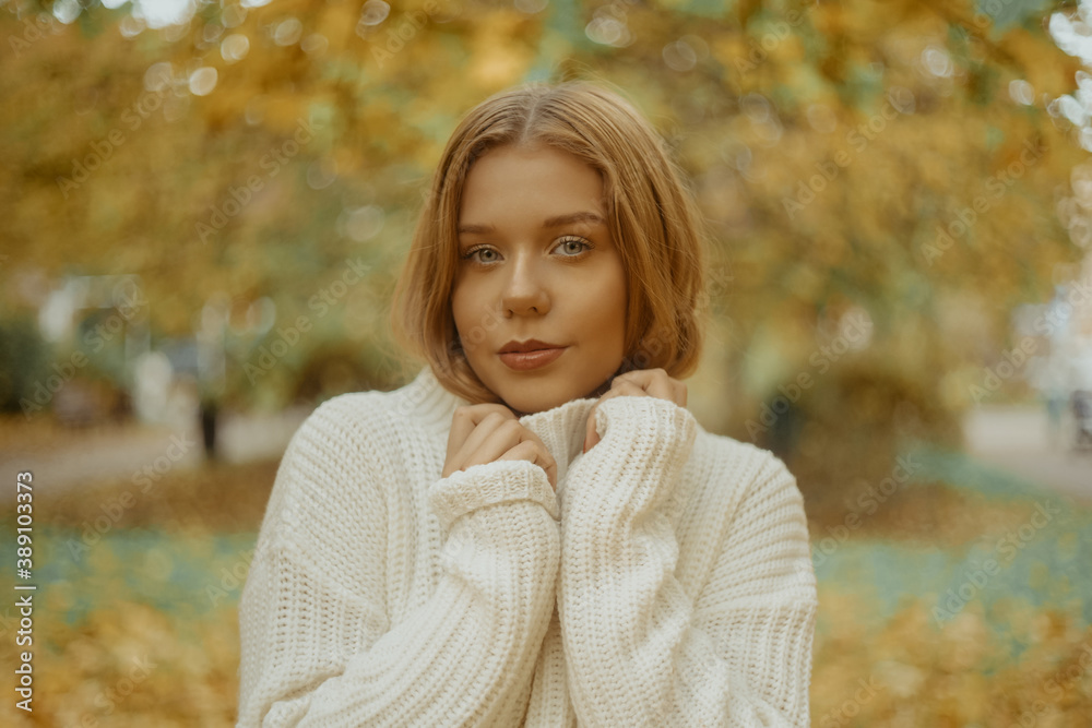 Photo portrait of a girl in a white sweater, the blonde holds the neck of the sweater and looks at the camera. Pretty woman in the park in autumn. Autumn fashion and style