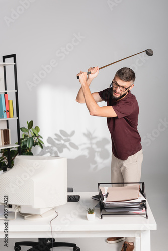 Angry businessman with golf club standing near computer and documents on blurred foreground in office