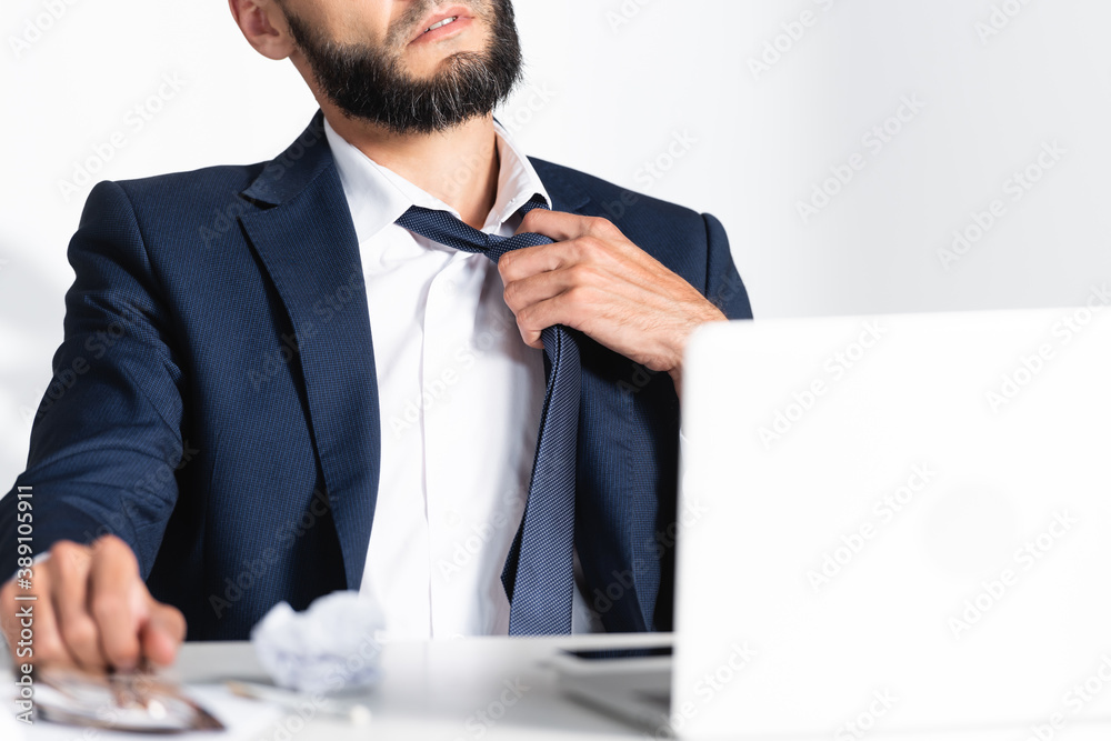 Cropped view of businessman touching tie while feeling hot near laptop on blurred foreground Cropped view of businessman touching tie while feeling hot near laptop on blurred foreground