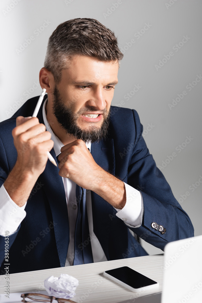 Tensed businessman holding broken pencil near smartphone, clumped paper and eyeglasses on blurred foreground Tensed businessman holding broken pencil near smartphone, clumped paper and eyeglasses on blurred foreground