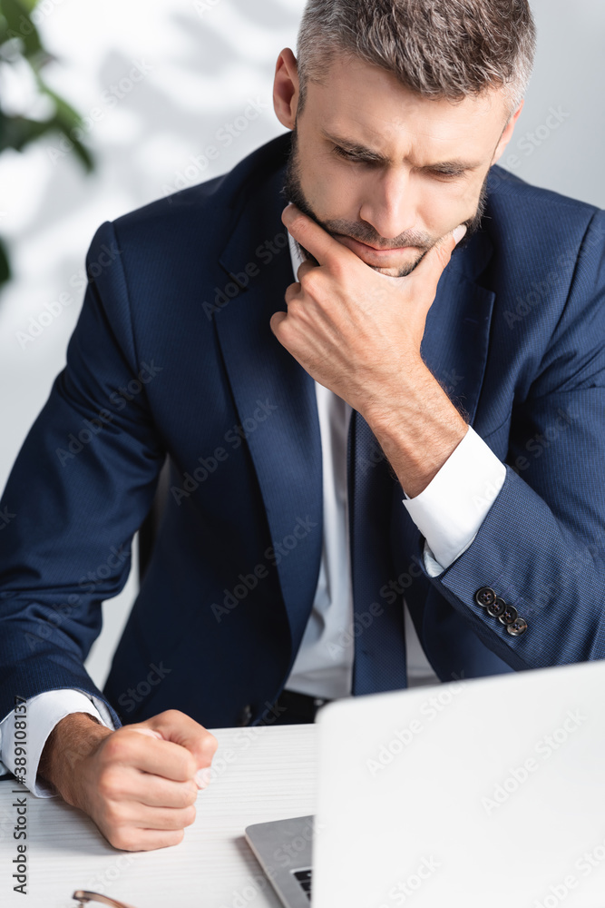 Pensive businessman with hand near chin sitting near laptop on blurred foreground Pensive businessman with hand near chin sitting near laptop on blurred foreground