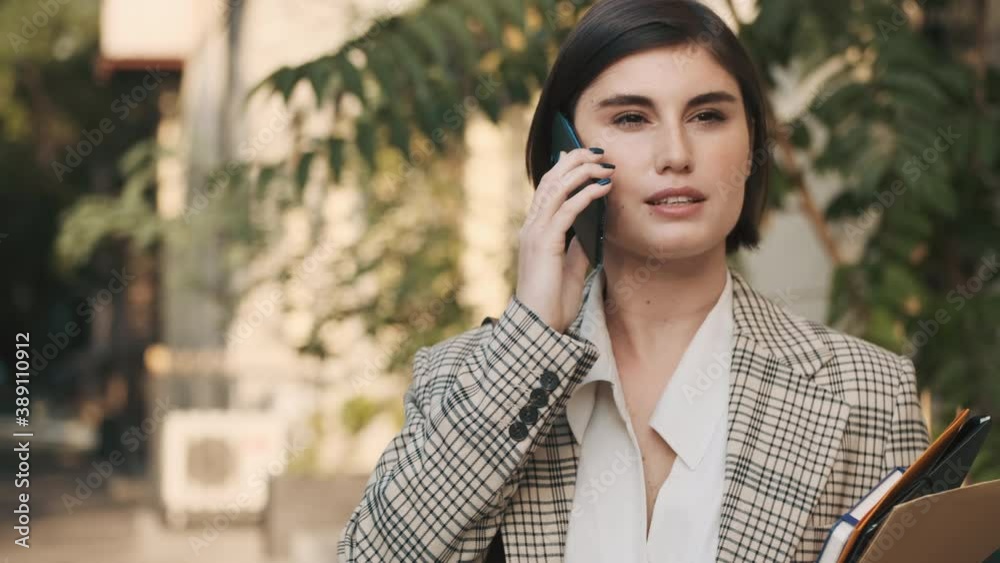 Tracking out shot of young businesswoman dressed in plaid blazer talking on smartphone waiting business meet on cozy city street. Modern estate agent waiting customers outdoor Tracking out shot of young businesswoman dressed in plaid blazer talking on smartphone waiting business meet on cozy city street. Modern estate agent waiting customers outdoor
