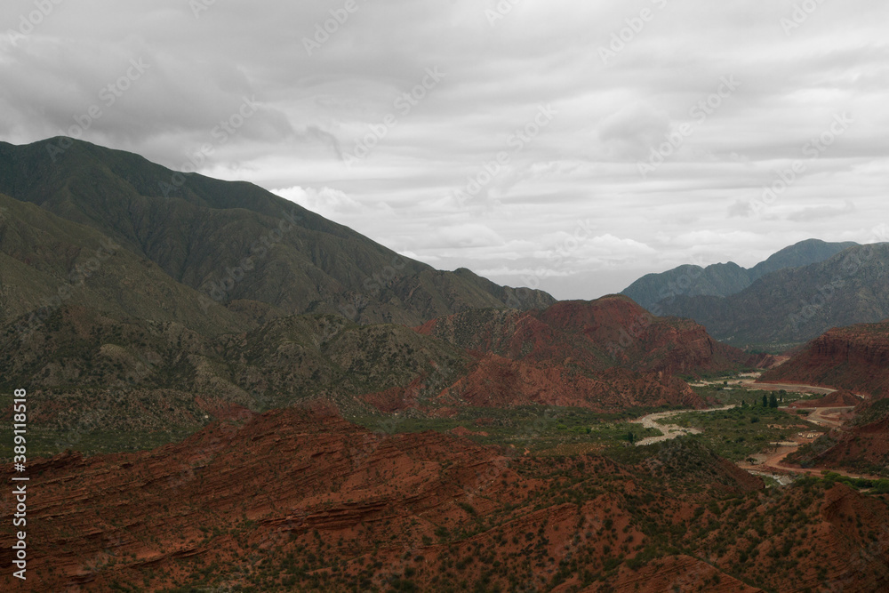 Geology. Panorama view of the red sandstone and rocky mountains, green forest and river flowing across the valley under a cloudy sky in Miranda Slope, La Rioja Argentina. 