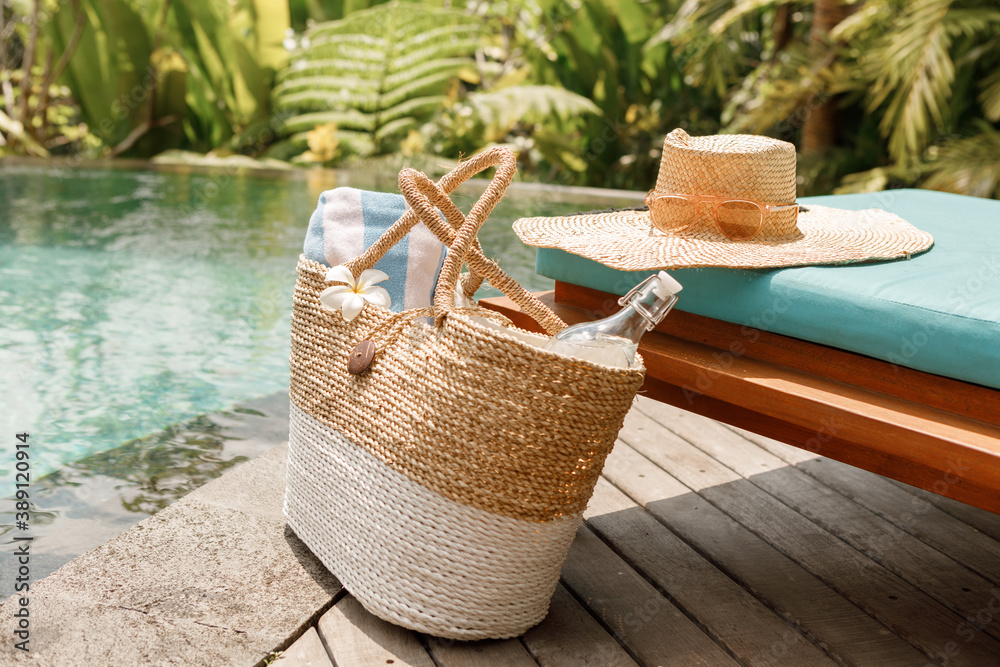 Summer beach bag with straw hat,towel,sunglasses and bottle of water near swimming pool