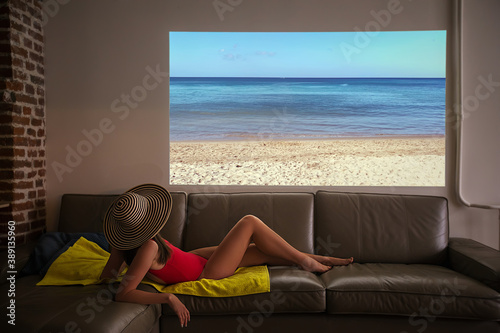 Woman pretends to sunbathe on a fake beach looking at the sea of a projector during a pandemic at home in an apartment