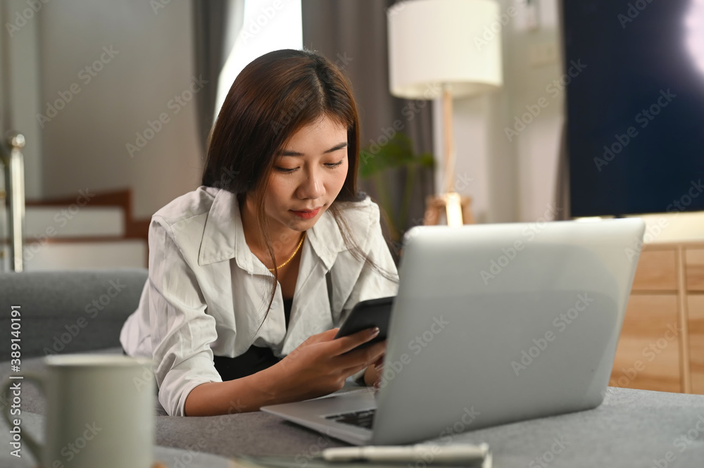 A young woman is relax on comfortable sofa in living room while use modern cellphone and laptop. A young woman is relax on comfortable sofa in living room while use modern cellphone and laptop.