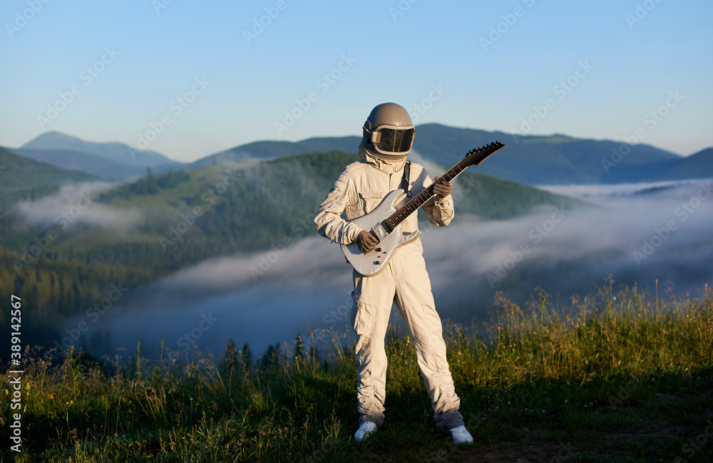 Naklejka premium Astronaut in white space suit and helmet playing guitar, standing on sunny green mountain glade in summer, morning fog rising up from the valley behind him. Concept of astronautics, music and nature.