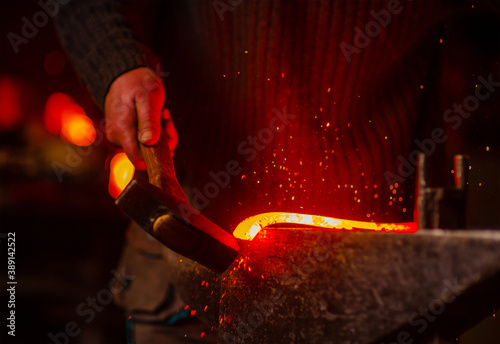 The blacksmith hits the red-hot workpiece in the forge with a hammer and glowing sparks fly in all directions