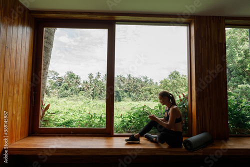 Young woman sit on the window in yoga studo with yoga mat and use mobile phone, lokk at the window with tropical view