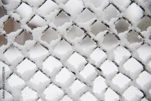 Snow lies on a metal fence. Snowflakes on a metal mesh. A photo with a shallow depth of field. Background image. Fallout of precipitation in the form of snow. Winter weather.