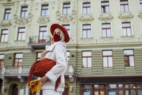 Outdoor fashion, lifestyle autumn portrait of elegant woman wearing trendy outfit with orange cloth protective face mask, hat, handbag, posing in street of European city. Copy, empty space for text  © Victoria Fox