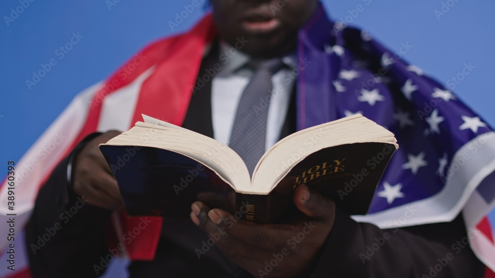 Φωτογραφία Close up, holy bible in the hands of black african american man in elegant suit with USA flag over the shoulders