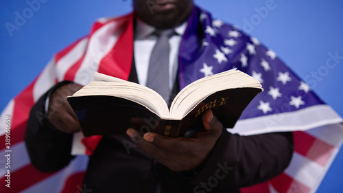 Fotografia, Obraz Close up, holy bible in the hands of black african american man in elegant suit with USA flag over the shoulders