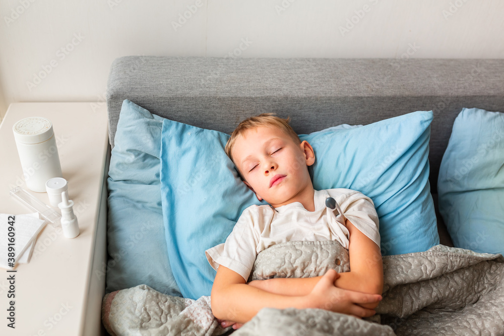 Sick little boy with high fever and headache laying in bed and holding thermometer. Stay at home during corona virus epidemic