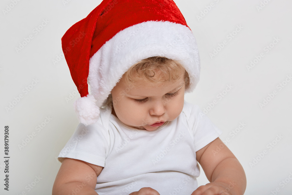 Cute baby wearing christmas clothes playing indoor, looking attentively at something in his hands, has blond wavy hair, has concentrated look, posing isolated over white background. Cute baby wearing christmas clothes playing indoor, looking attentively at something in his hands, has blond wavy hair, has concentrated look, posing isolated over white background.