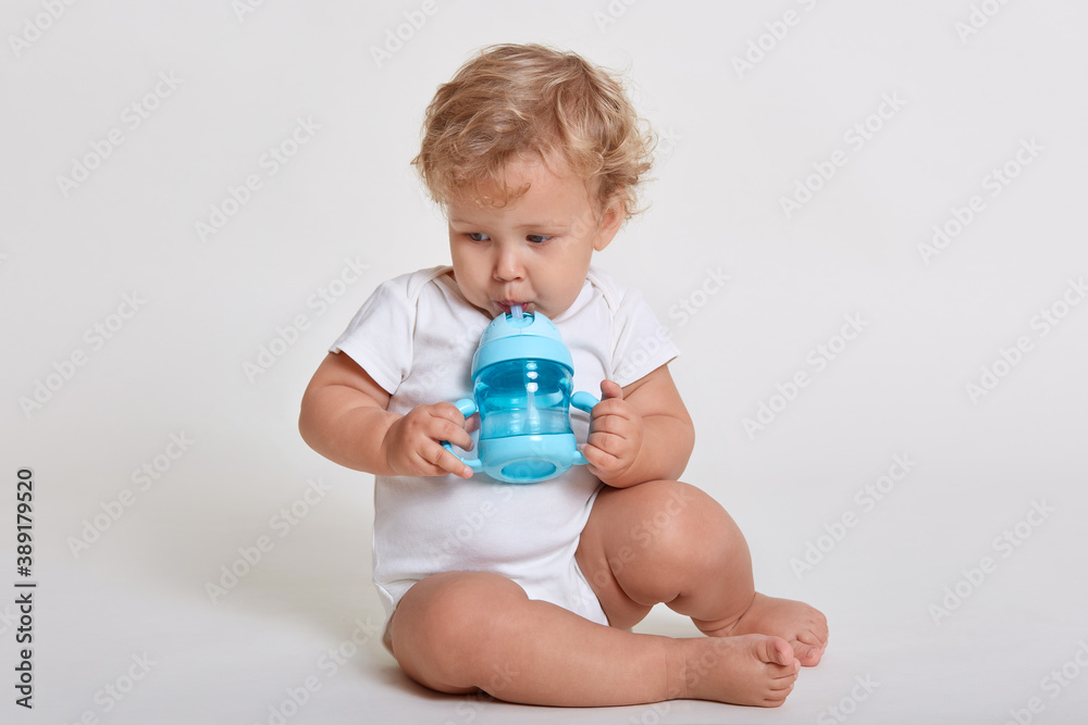 Little cute concentrated kid looking away while drinking water from baby cup, child with blonde curly hair sitting on floor, wearing body suit, posing isolated over white background. Little cute concentrated kid looking away while drinking water from baby cup, child with blonde curly hair sitting on floor, wearing body suit, posing isolated over white background.